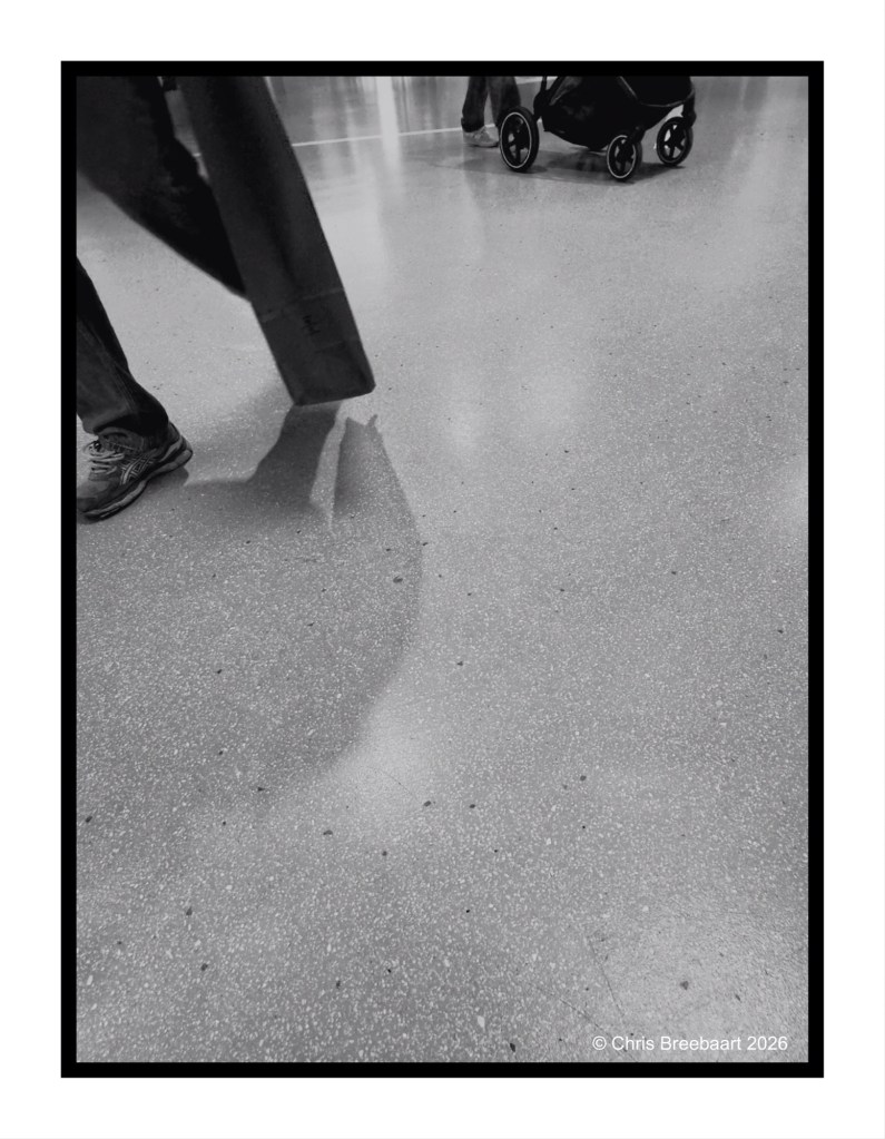 Black and white image of a floor with shadows, showcasing the legs of people walking and a stroller in the background.