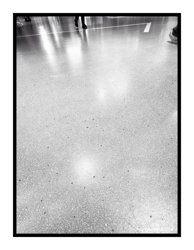 A black and white image of a smooth, speckled floor with a faint reflection, showing the lower part of legs and feet of people in the background.