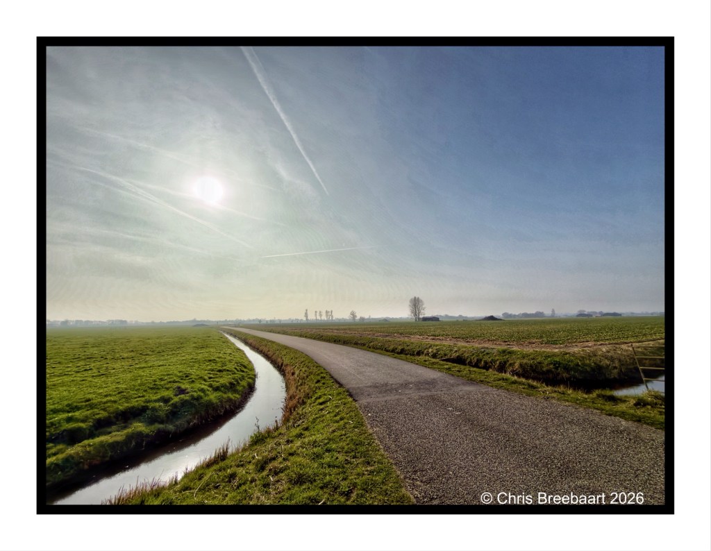 A scenic view of a winding path next to a small canal, surrounded by lush green fields under a clear sky with sun and wispy clouds.