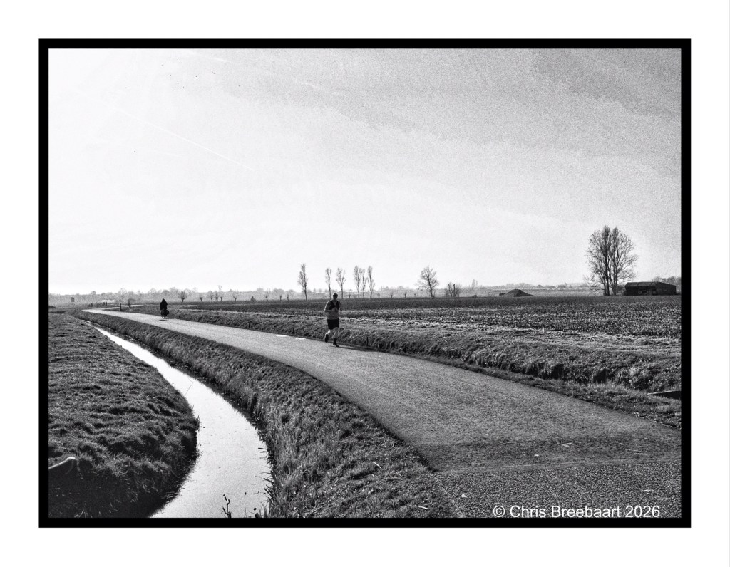 A black and white image of a winding road through a rural landscape, with two people walking along the path and a small stream running alongside. The horizon is lined with trees and fields.