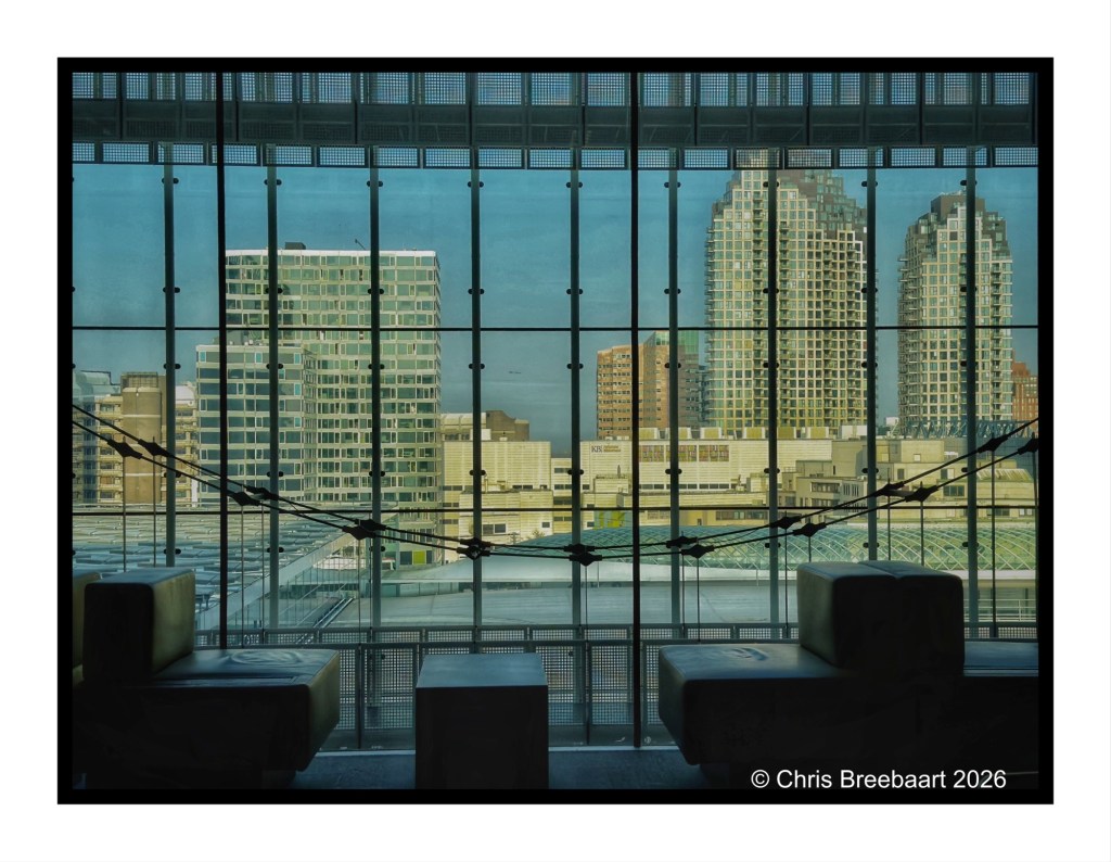 A modern urban skyline view through large glass panels, featuring various high-rise buildings and city architecture, with seating visible in the foreground.