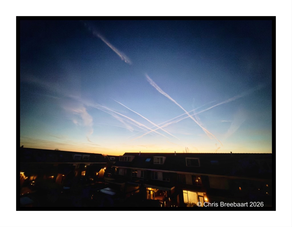 A twilight sky filled with streaks of contrails, with silhouettes of houses in the foreground.