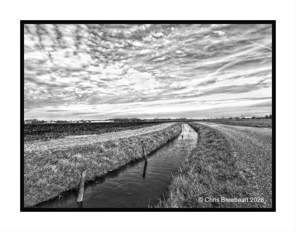 A monochrome landscape featuring a winding canal bordered by grassy banks, under a cloudy sky. Fields stretch out in the background.