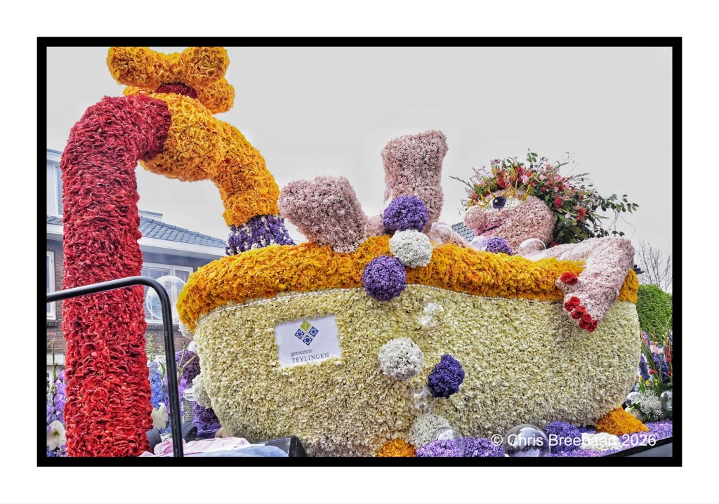 A large floral float featuring a bathtub filled with colorful blooms, adorned with a figure relaxing inside. The float is decorated with various flowers in vibrant hues, including red, yellow, and purple, enhancing its festive appearance.