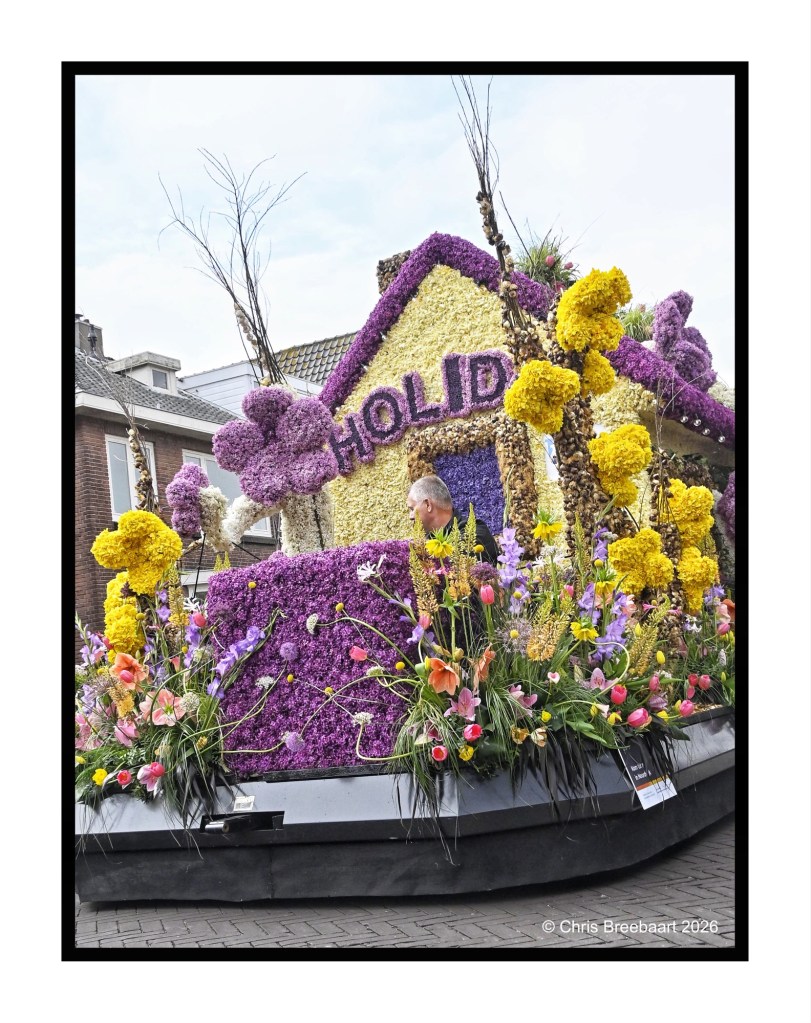 A colorful floral float designed to resemble a house with the word 'HOLDI' prominently displayed, surrounded by various types of flowers and greenery at a festive event.