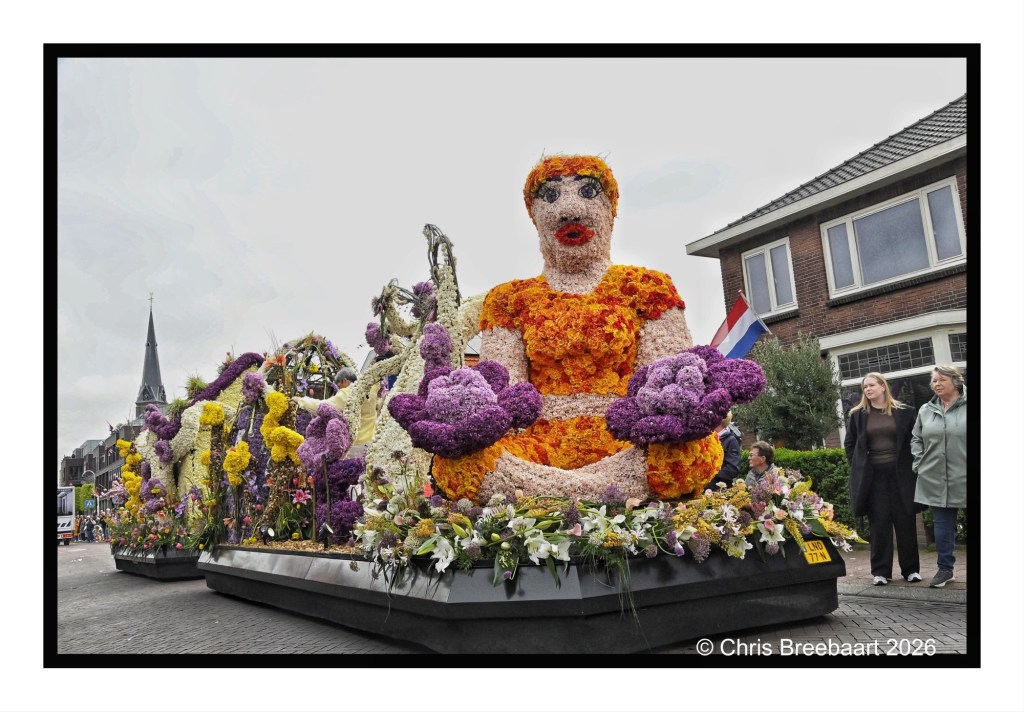 A float decorated with a large floral sculpture of a seated figure holds colorful flowers, set against a backdrop of onlookers and buildings during a parade.