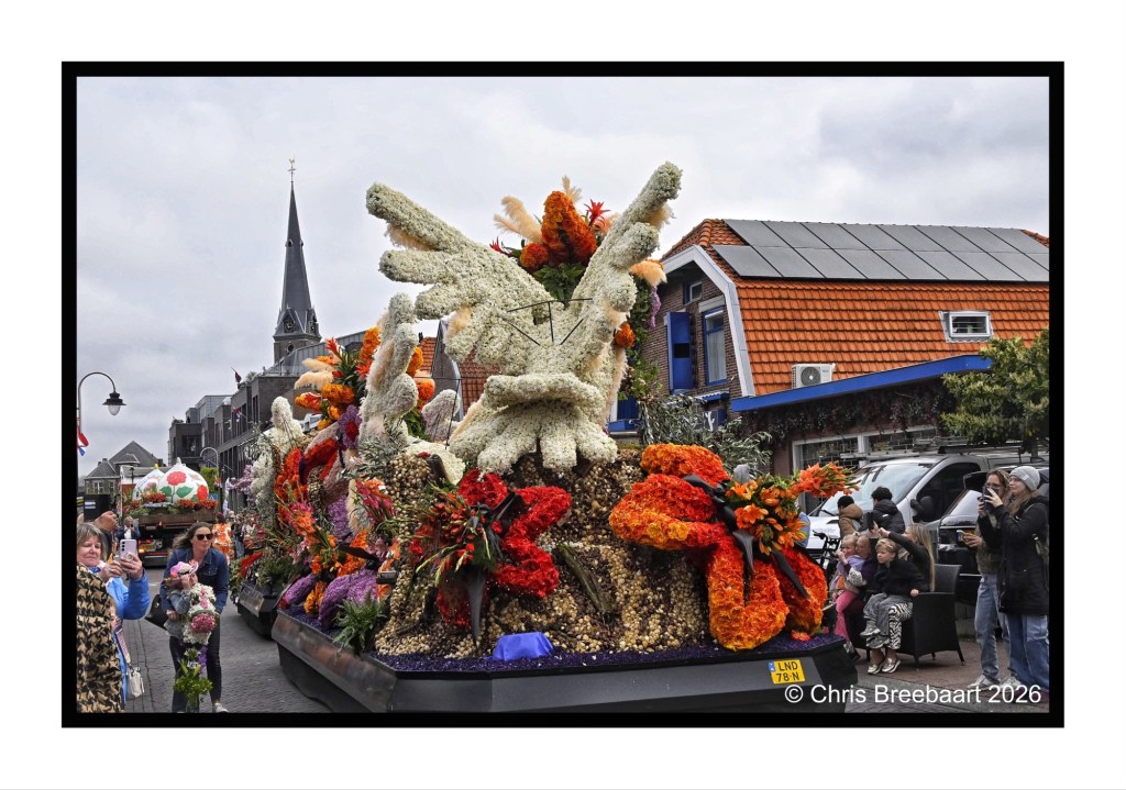 A colorful floral float adorned with intricate arrangements of flowers, featuring a prominent hand sculpture, showcased during a parade in a town square, with spectators gathered around.