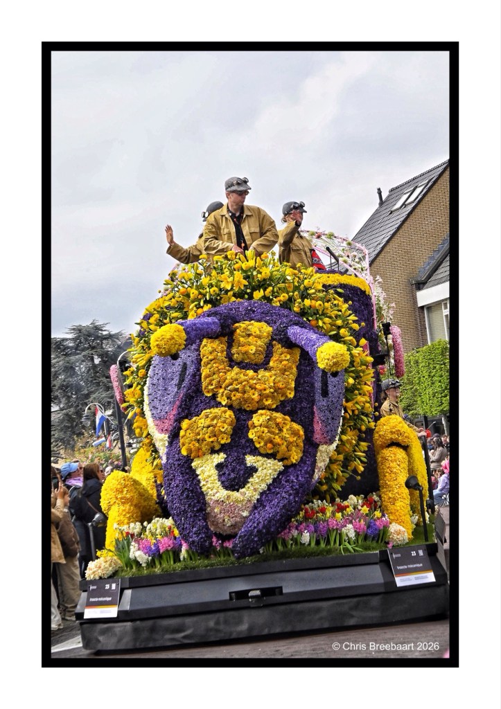 A floral float adorned with colorful flowers, featuring two individuals dressed in light-colored jackets and caps, parading down a street during a festival. The float showcases vibrant floral designs and is surrounded by spectators.