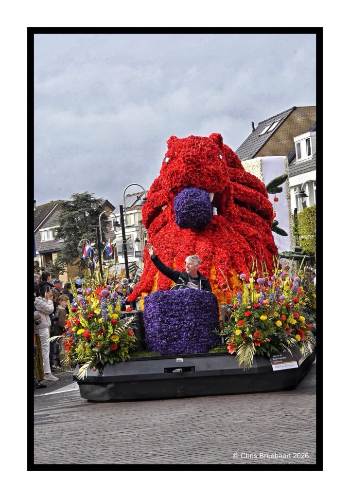 A float decorated with vibrant flowers in the shape of a large octopus, featuring a speaker at a podium surrounded by floral arrangements during a parade.