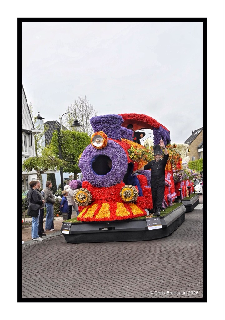 A colorful flower-covered float designed as a train, featuring a person in a black outfit waving. In the background, onlookers are gathered along the street during a parade.