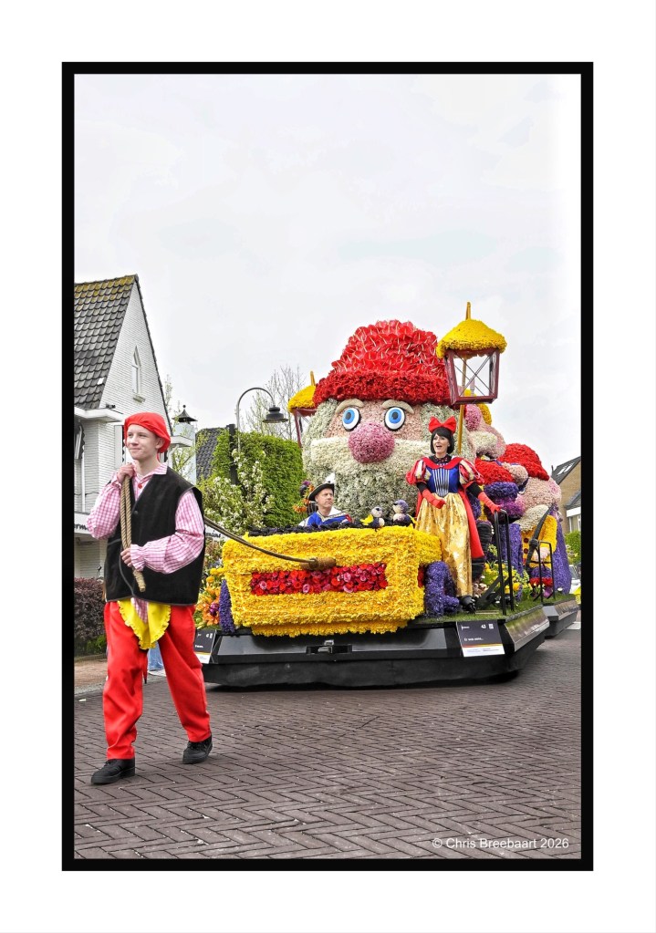 A colorful parade float decorated with flowers, featuring a whimsical character with large eyes and a red hat. In the foreground, a performer in a striped shirt and red pants walks alongside the float, while others in costume are seated on it.