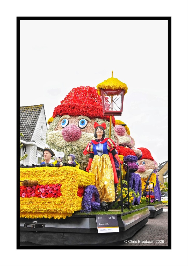 A colorful parade float featuring a large cartoonish figure with a red hat and oversized eyes, surrounded by vibrant floral decorations. A woman dressed as Snow White stands on the float, with a lantern beside her, while another participant sits nearby.
