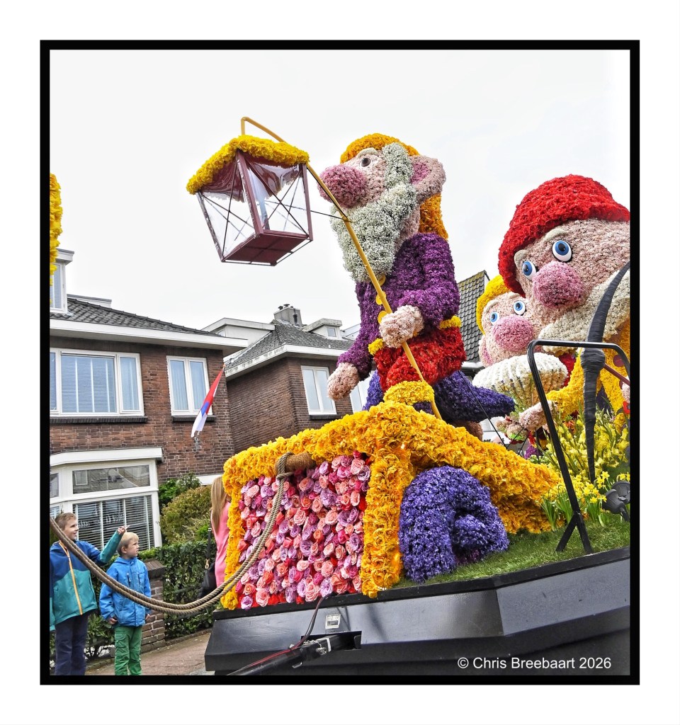 A vibrant float decorated with colorful flowers featuring animated dwarf-like figures, including one holding a lantern, in a parade setting. Two children are watching in the foreground.