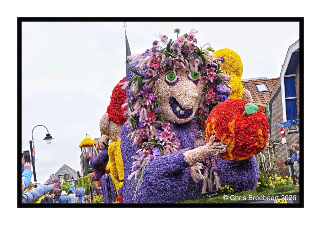 A large floral parade float depicting a whimsical character with green eyes and a colorful flower headdress, holding a large orange ball, surrounded by onlookers in a festive outdoor setting.