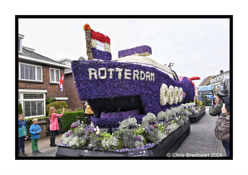 A large floral float shaped like a boat, decorated with vibrant flowers and the word 'ROTTERDAM' prominently displayed. Several spectators, including children, admire the float in a street lined with houses.