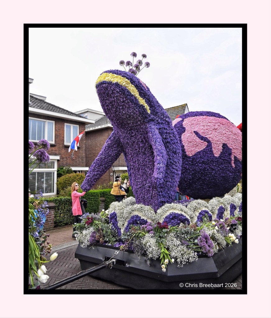 A large floral sculpture of a purple creature, resembling a snail, adorned with flowers and surrounded by greenery. People are interacting with the sculpture during a festive event.