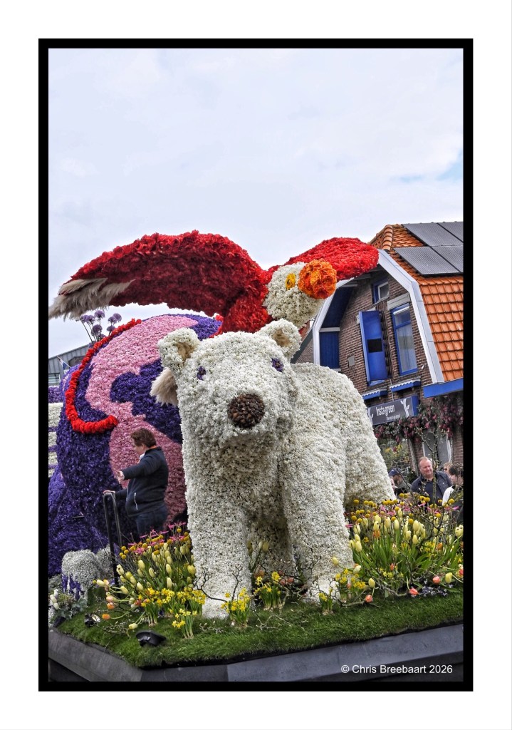 A large floral sculpture of a polar bear with purple and red accents, surrounded by colorful flowers and a crowd of people in the background.