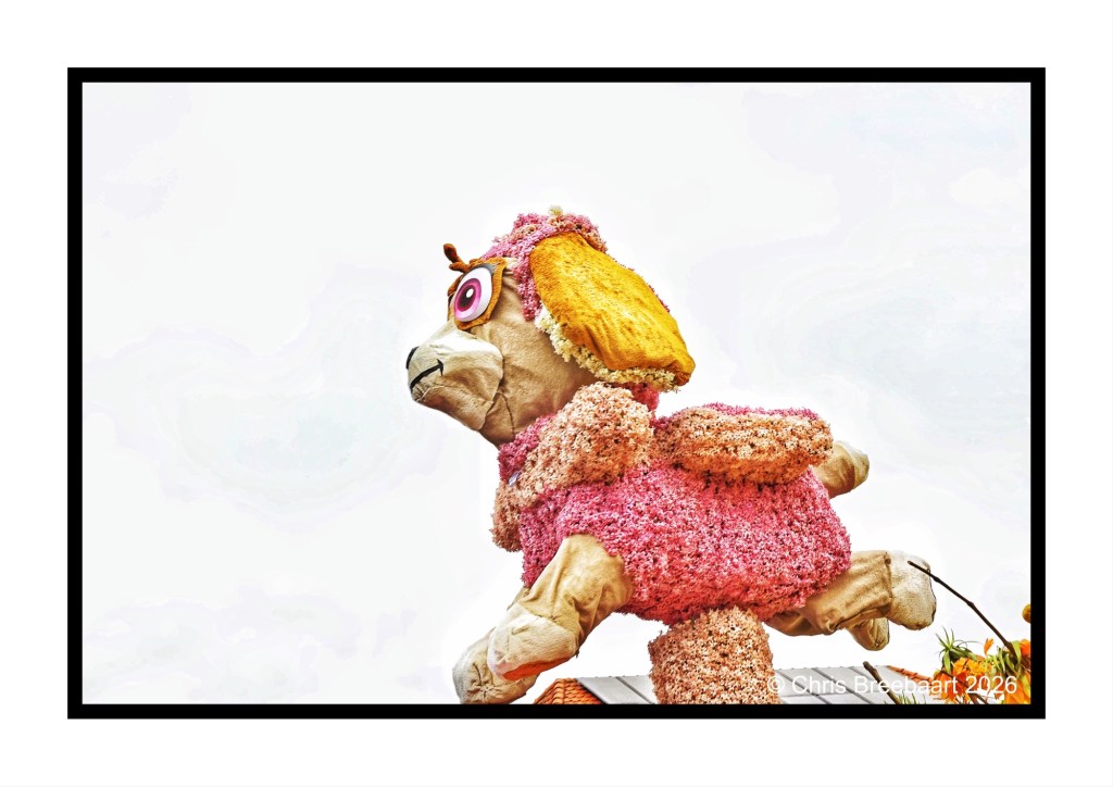 A colorful floral float featuring a large, stylized dog sculpture with big eyes and a pink coat, positioned against a light sky.