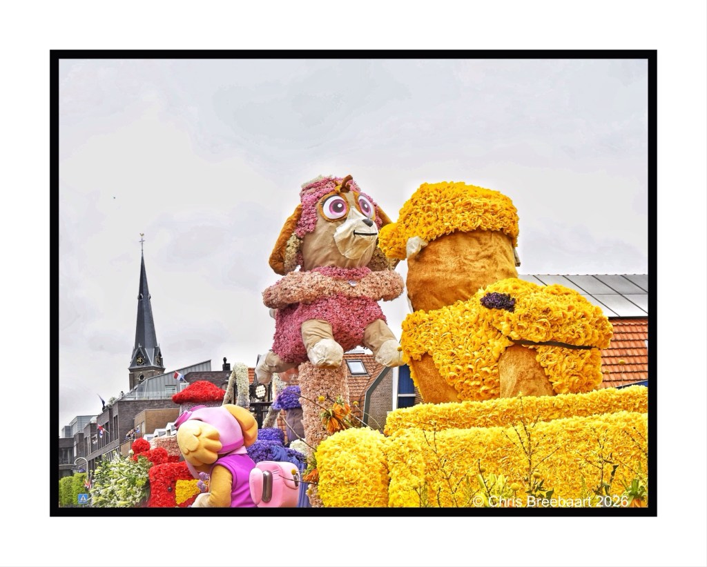 Floats decorated with vibrant flowers featuring large animal characters at a festival, with a church spire in the background.