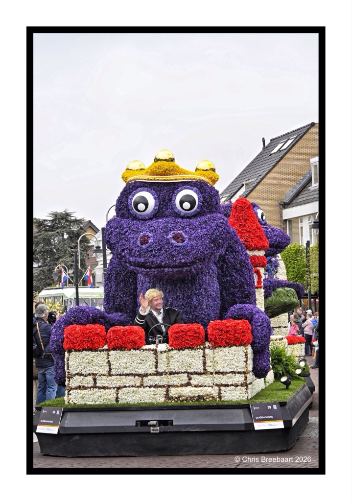 A colorful float featuring a large purple creature wearing a crown, surrounded by floral decorations, with a man waving from the front.
