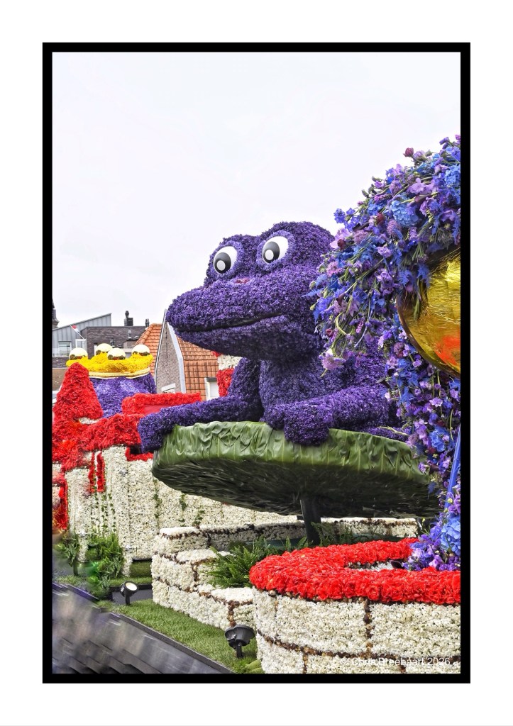 A large purple frog sculpture made of flowers sits on a green lily pad, surrounded by colorful floral decorations in a garden setting.