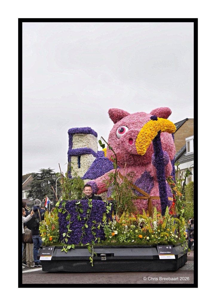 A large colorful float in a parade featuring a pink and purple floral arrangement shaped like an animal, with a person seated in front surrounded by greenery and flowers.