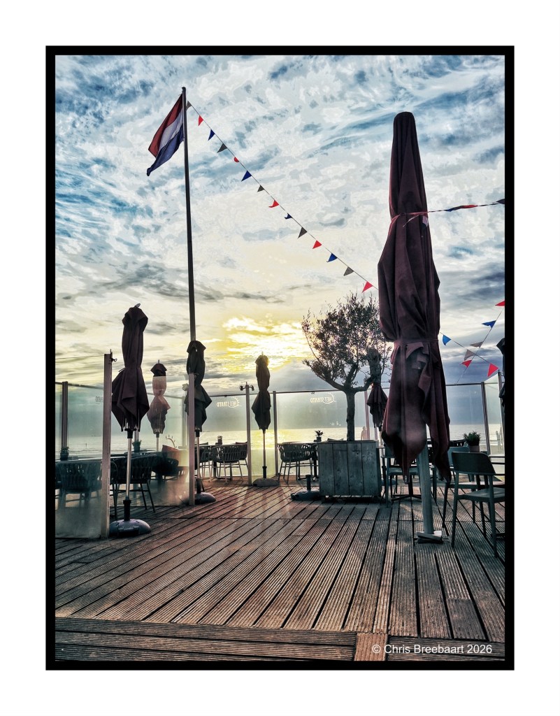 Outdoor dining area with tables and chairs, featuring closed umbrellas; a flag and colorful bunting visible against a sunset sky with clouds.