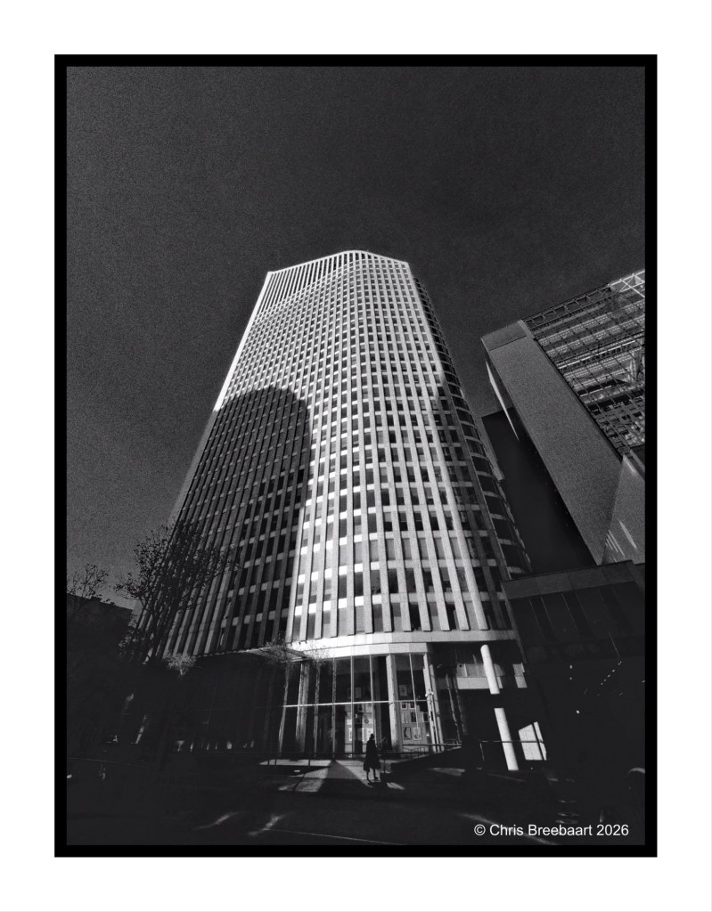 A black and white image of a modern skyscraper viewed from a low angle, showcasing its towering height and unique architectural features, with shadows cast on the facade.
