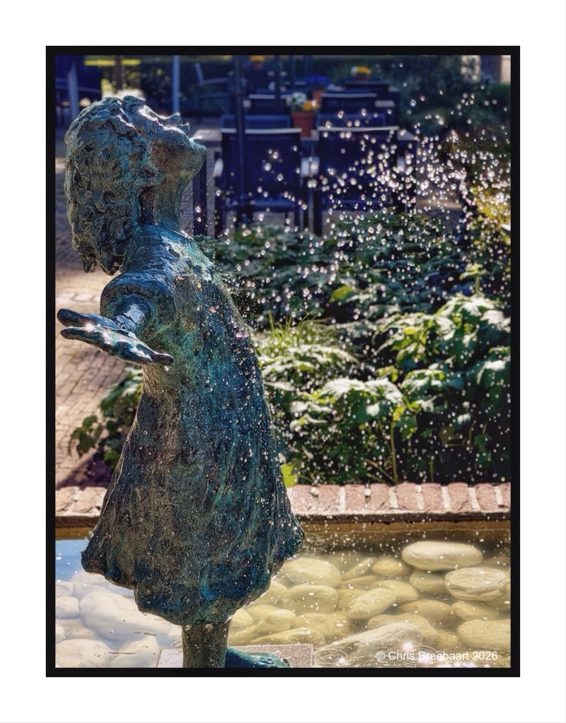 A bronze statue of a joyful child with arms outstretched, standing near a fountain with water droplets shimmering in the sunlight, surrounded by greenery.