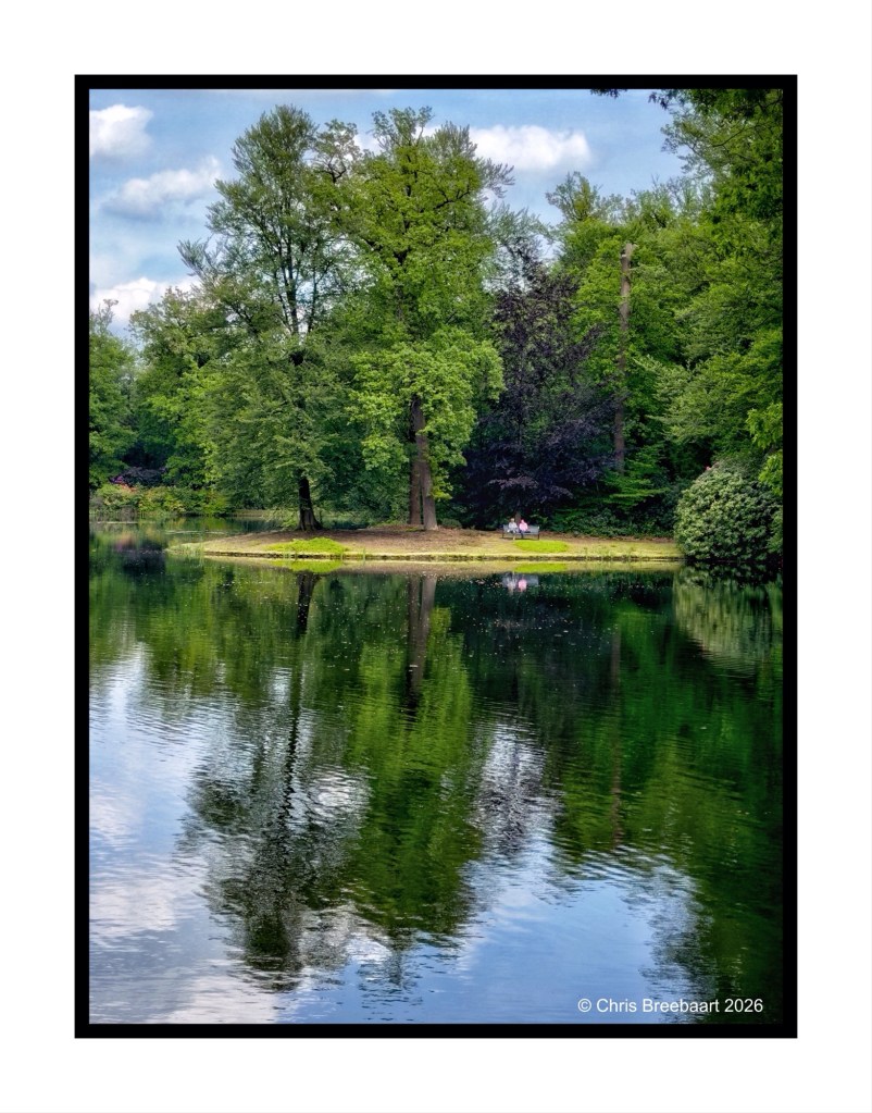 A serene pond surrounded by lush green trees, reflecting the vibrant foliage and blue sky, with two people seated on a small island by the water's edge.