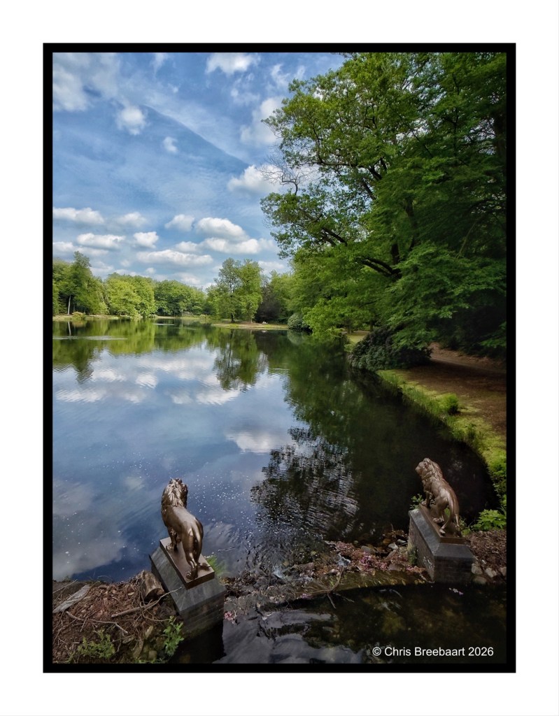 A serene park scene featuring a reflective lake surrounded by lush greenery, with two lion statues positioned at the water's edge.