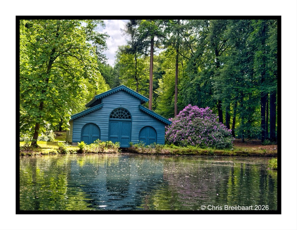 A serene blue wooden boathouse surrounded by lush greenery and a flowering bush, reflecting in a calm pond.