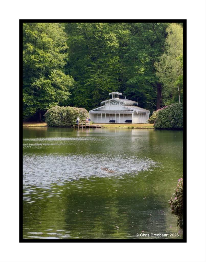 A serene lake surrounded by lush greenery, featuring a white gazebo-like structure by the water and two people near a wooden dock.