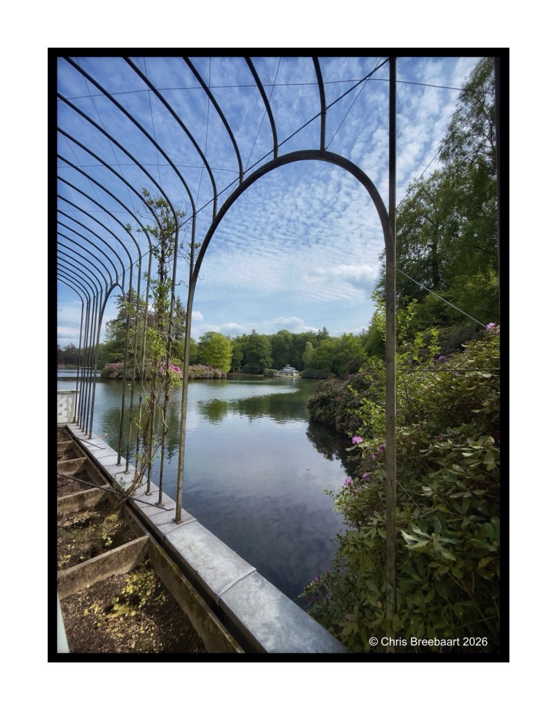 A scenic view of a calm lake framed by a decorative metal arch, with lush greenery and colorful flowers along the banks, under a partly cloudy sky.