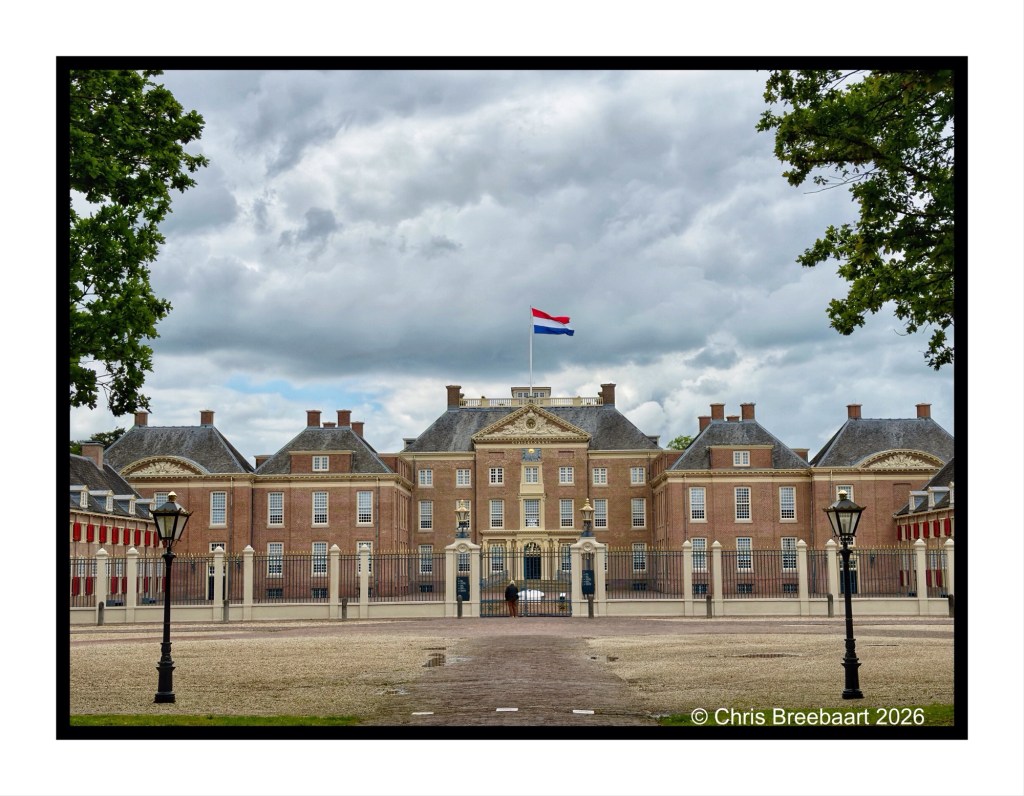 A historic building with a large facade, featuring multiple windows and a Dutch flag flying atop. The surrounding area includes a cobblestone pathway and decorative lamp posts, and the sky is filled with clouds.