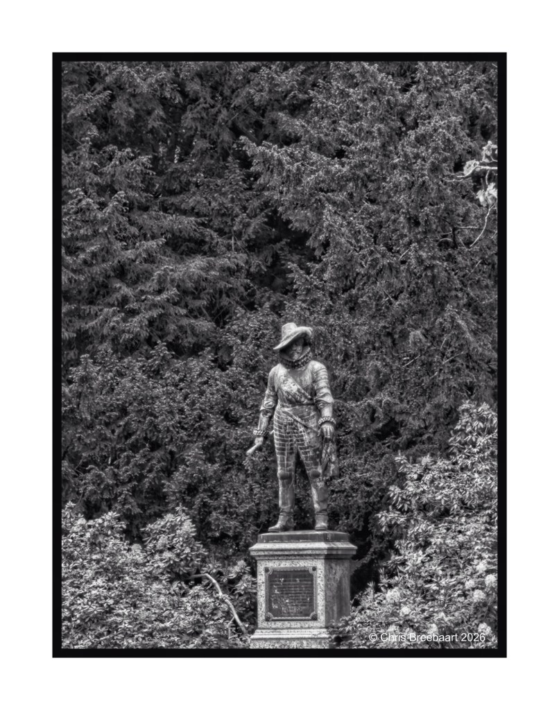 A black and white photograph of a statue depicting a standing figure in historical attire, set against a backdrop of dense foliage.