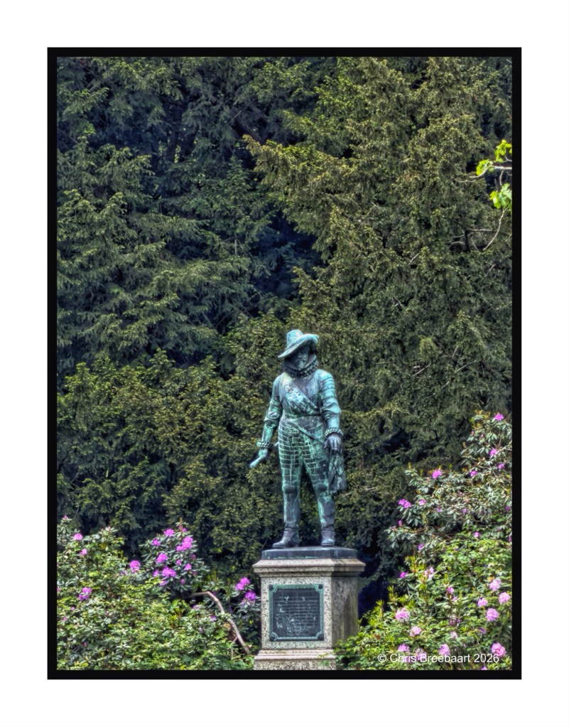A bronze statue of a historical figure standing on a pedestal, surrounded by vibrant green trees and flowering plants.