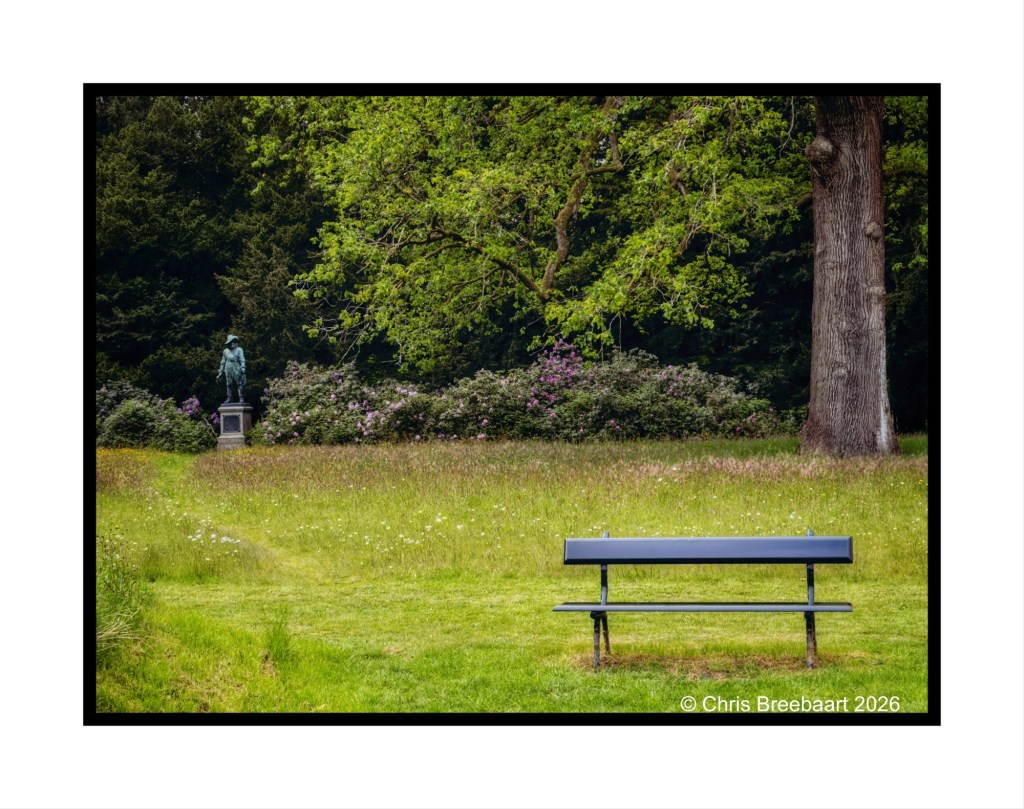 A tranquil park scene featuring a black bench in the foreground, a statue behind the bench surrounded by lush greenery and flowering bushes.
