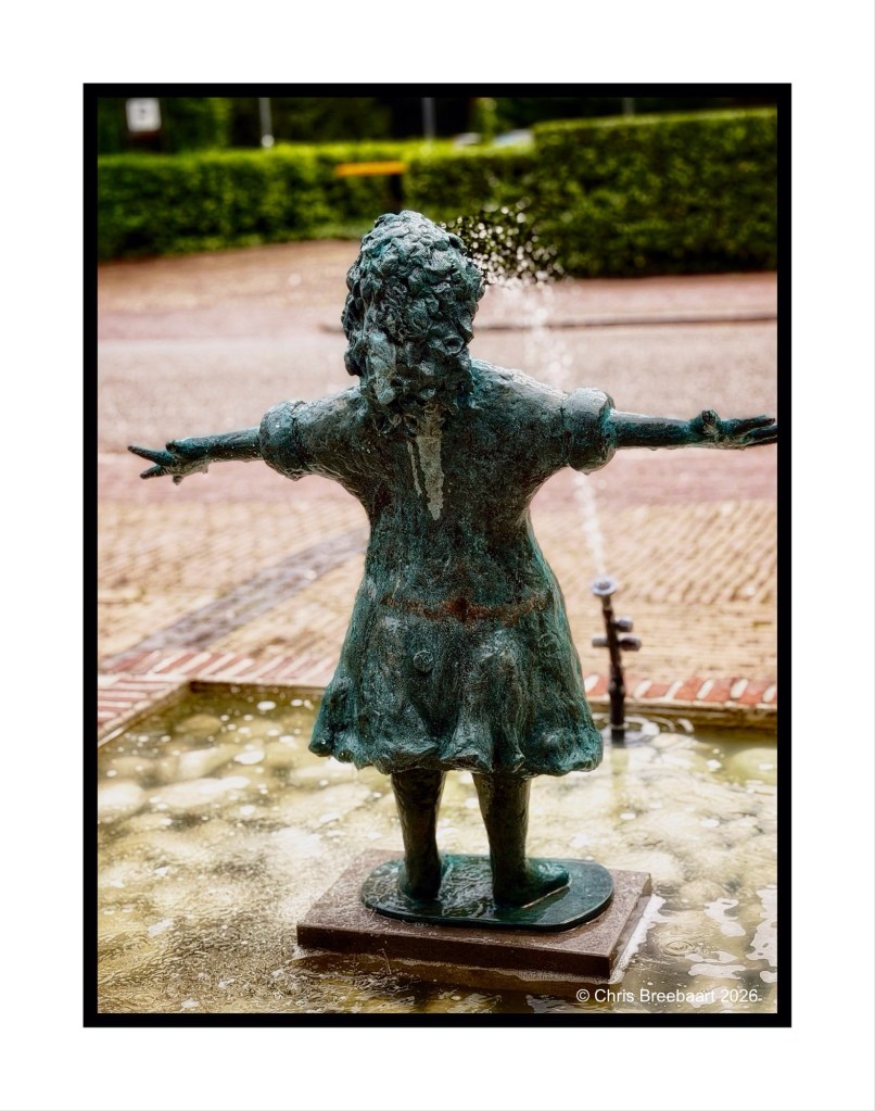 A bronze statue of a girl with curly hair, standing with arms outstretched, in a shallow water fountain.
