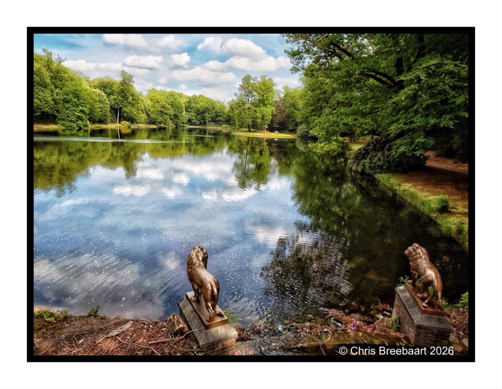 A tranquil lake surrounded by lush green trees, featuring two lion statues on stone pedestals at the foreground, reflecting clouds and foliage in the water.