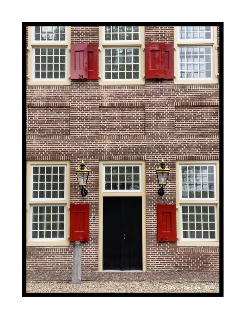 A brick building facade featuring large windows with red shutters, a black door in the center, and two ornate lanterns on either side.