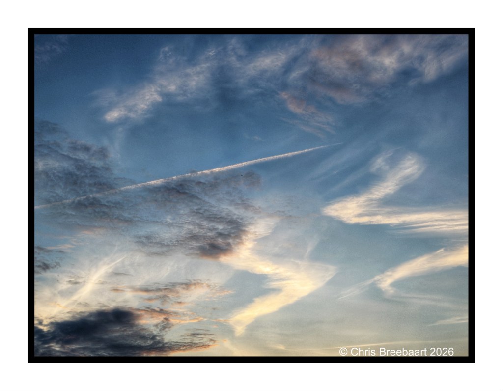 A picturesque sky filled with soft, wispy clouds against a blue backdrop, with a subtle sunset glow reflecting off the clouds and a faint jet trail crossing the scene.