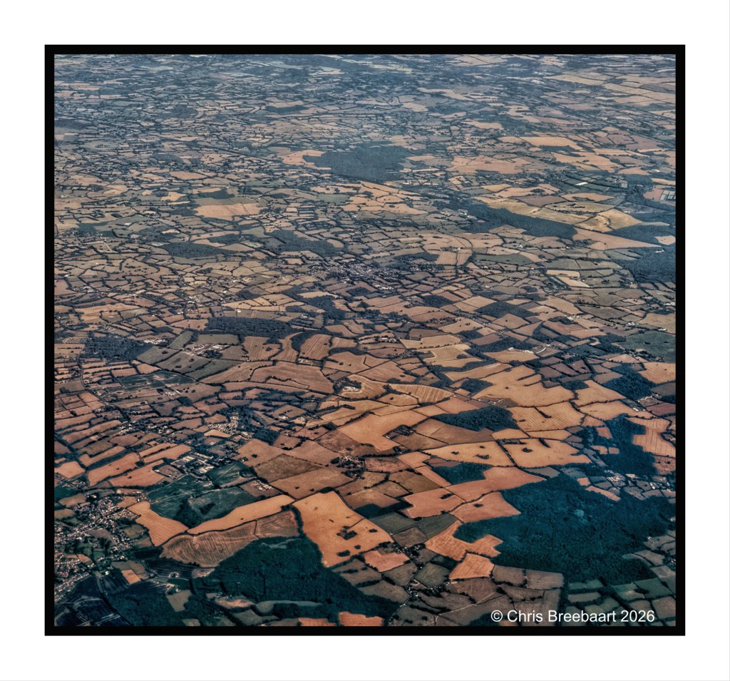 Aerial view of patchwork agricultural fields with varying shades of brown and green, interspersed with small settlements and clusters of trees.