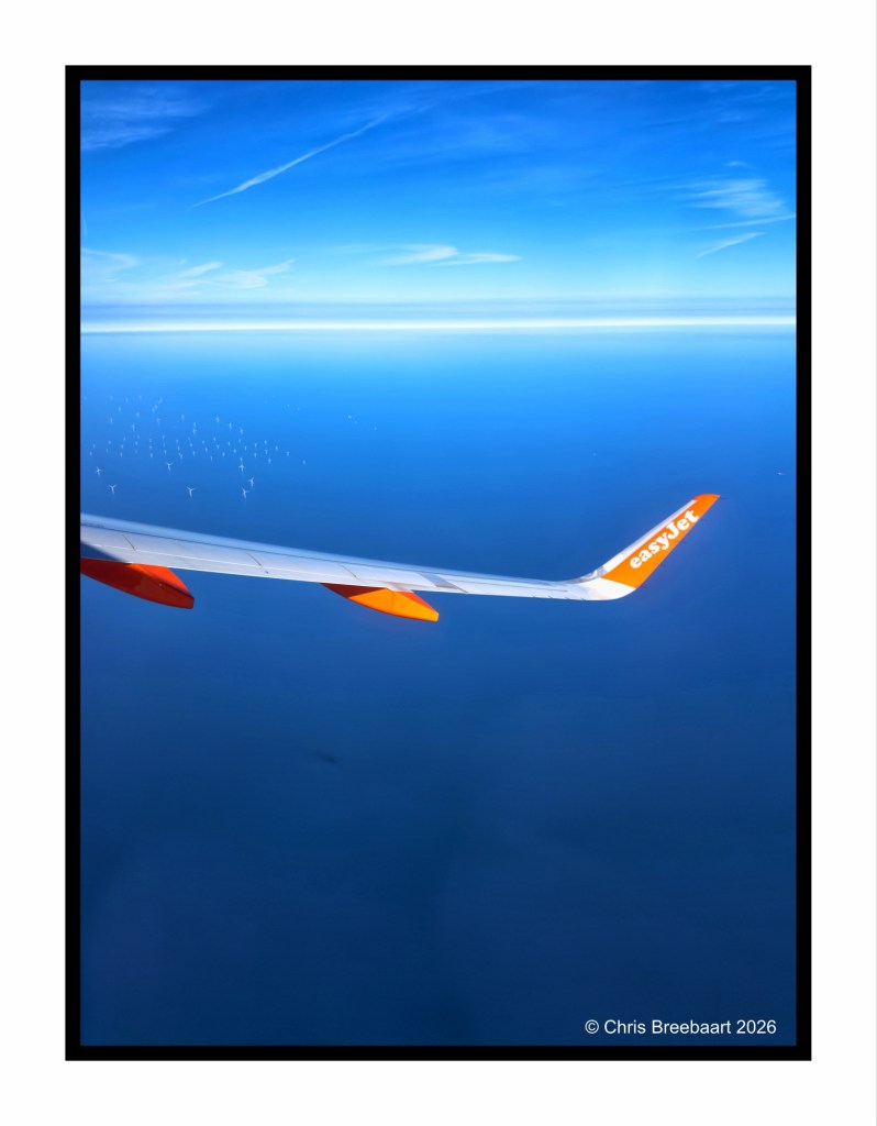 View from an airplane window showing the wing of an easyJet aircraft against a blue sky and ocean, with wind turbines visible in the distance.
