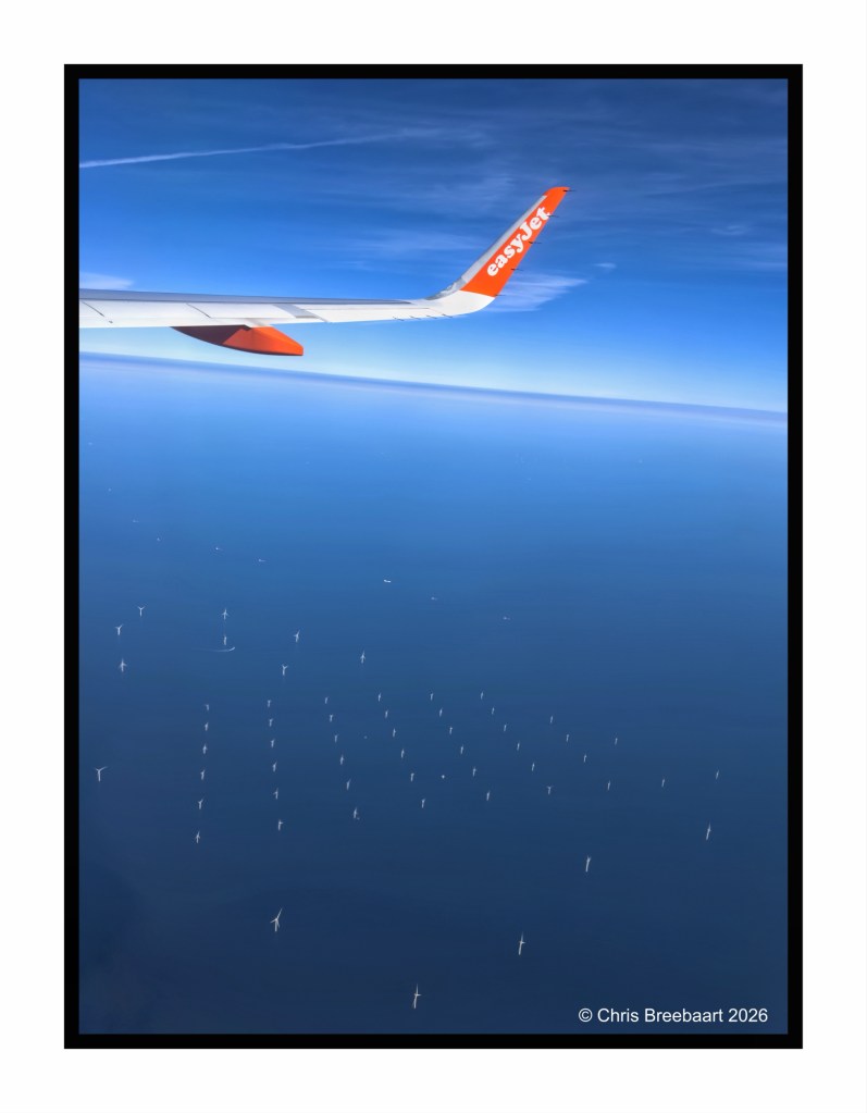 Aerial view of an airplane wing with the easyJet logo, overlooking a blue ocean populated with numerous wind turbines in the distance.
