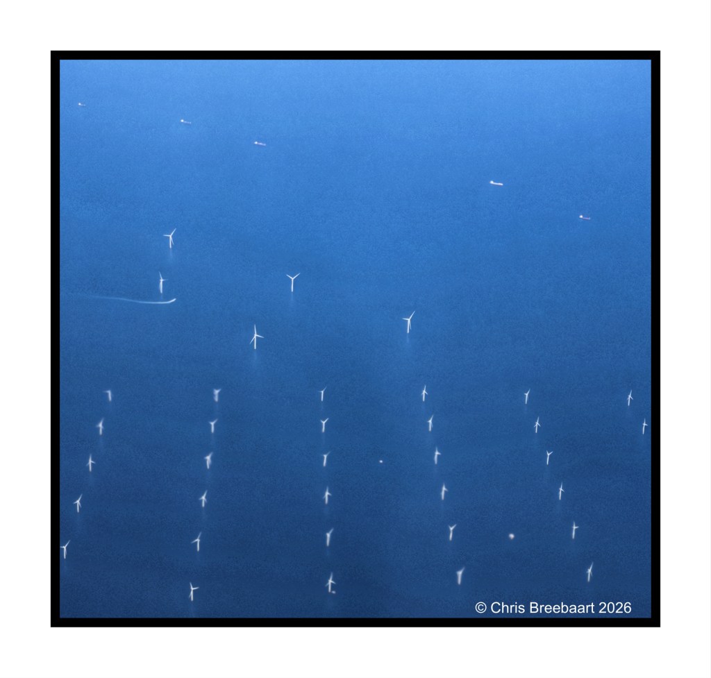 Aerial view of several wind turbines in a blue ocean, with faint silhouettes of boats in the background.