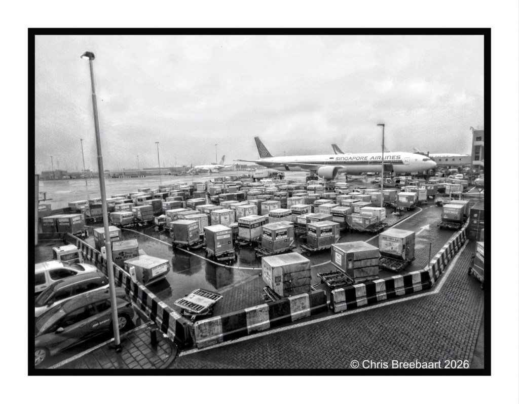 A black and white image of an airport cargo area showing numerous shipping containers and a Singapore Airlines airplane in the background.