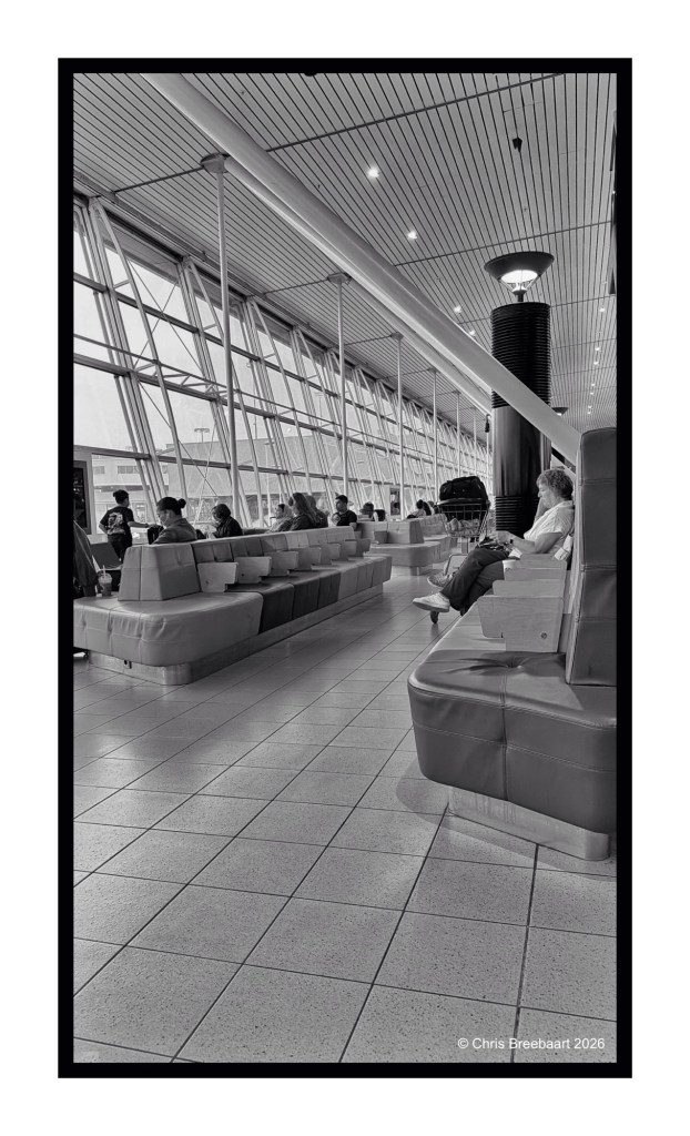 A modern airport terminal interior showcasing spacious seating areas with passengers waiting. The photo is in black and white, highlighting the architectural elements like large glass windows and stylish lighting fixtures.