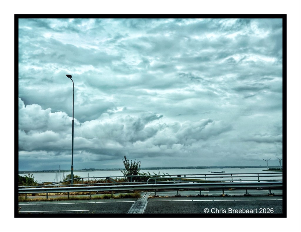 A scenic view of a cloudy sky over a body of water, with a highway guardrail in the foreground and a streetlamp on the left.