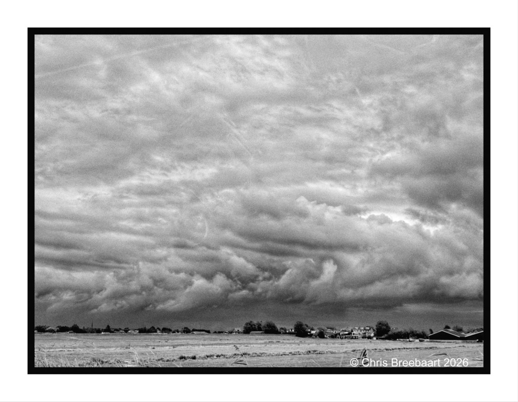 A black and white photograph of a dramatic sky filled with textured clouds, overlooking a rural landscape with distant houses and trees.